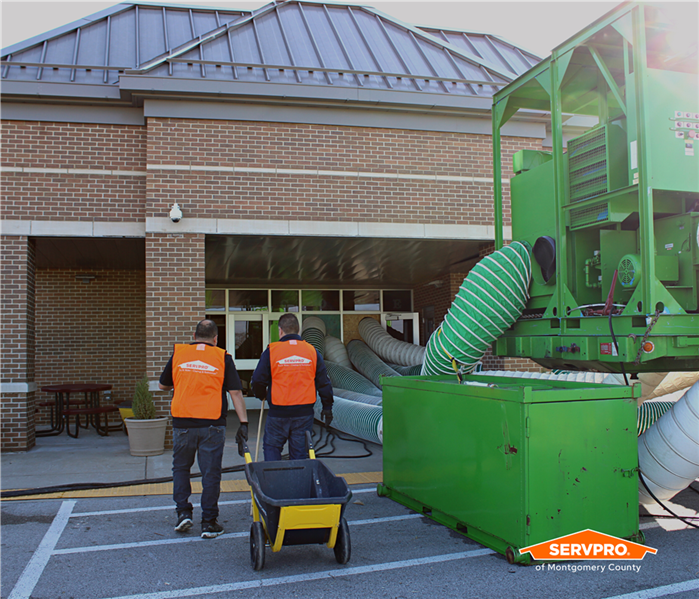Two SERVPRO of Montgomery County employees in orange vests walking into a school next to a big, green desiccant dehumidifier 